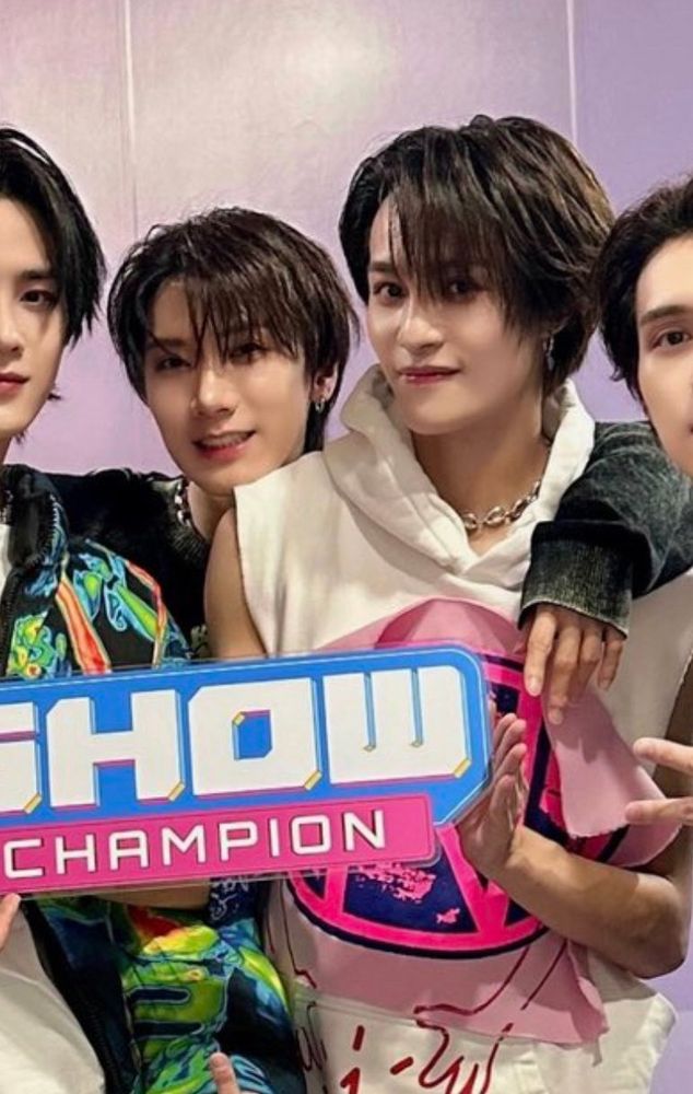 Ten and Yangyang in a group picture, holding a "Show champion" sign. Ten is standing behind Yangyang with his arm around his shoulder. 
They're both smiling and both have dark brown hair and Yangyang is wearing a sleeveless white hoodie.