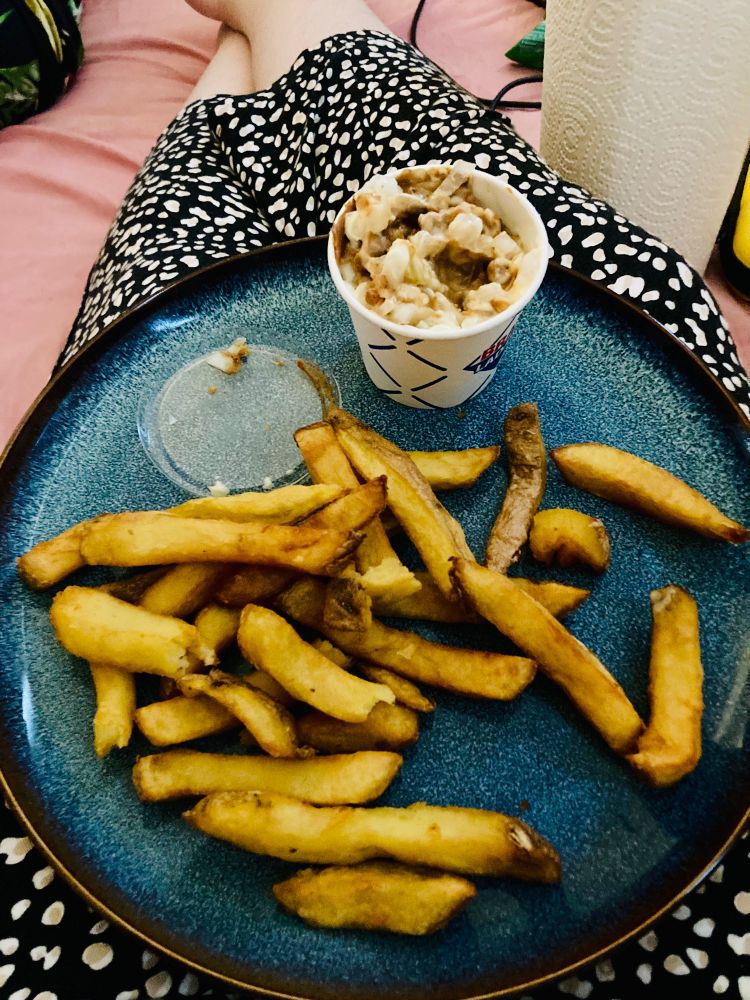 Een rond groen/blauw bord met daarop patatjes van Bran met een bekertje pindasaus/mayo/uitjes. Bord staat op mijn benen die languit op bed liggen.