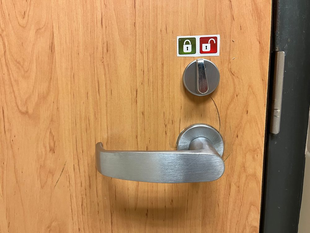 A door handle and lock with a color-coded label for the locked and unlocked position of the bolt lock. The “locked” graphic has a green background while the “unlocked” graphic has a red background.