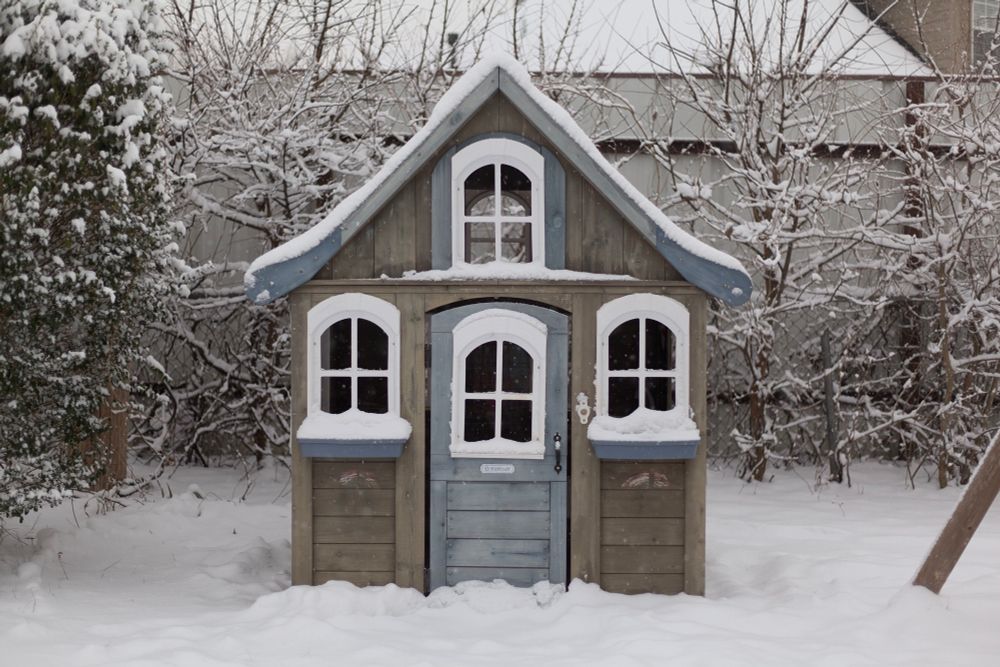 A play house for children, covered in a light dusting of snow. The front of the house features three windows in a triangular formation, with a windowed door in the center. The house was made by KidKraft and the house model is named Forestview II.