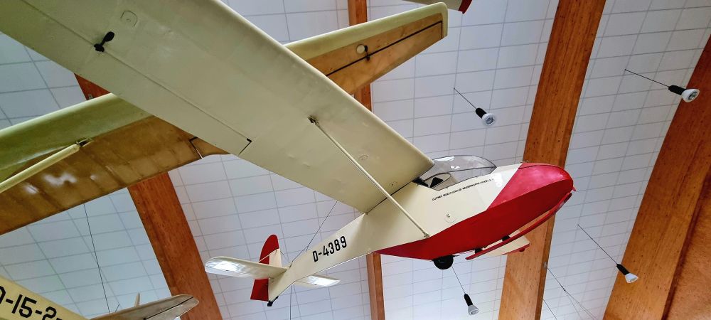 In einer großen Halle sind an der Decke verschiedene Segelflugzeuge aufgehängt. In der Bildmitte ein Model in weiß mit roten Akzenten, z.B. an der "Schnauze" und an der Heckflosse. 