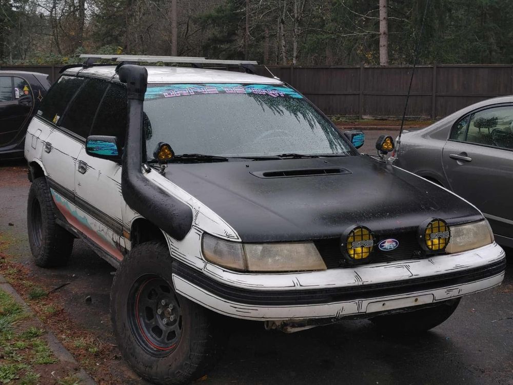 A white 90s Subaru legacy wagon lifted on steelies and running a snorkel exhaust and black hood, sporting a 90s wave style banner on the windshield