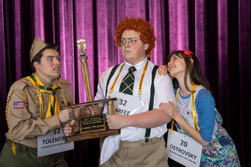 Photo from the 24th Annual Putnam County Spelling Bee. Three students competing in a spelling bee are fighting over a trophy on a theatre stage. 