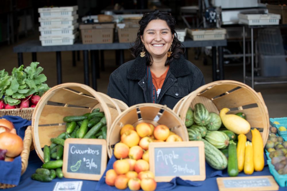 A young woman smiles while standing behind a table with produce for sale at SRJC's Shone Farm. 