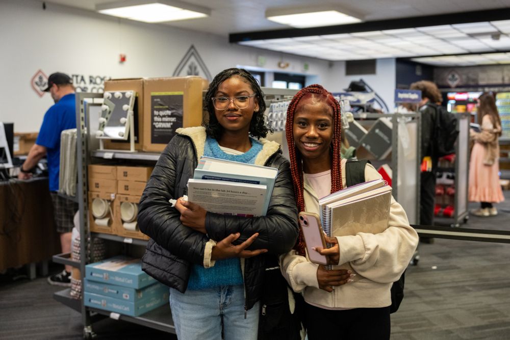 Two Black students are smiling at the camera while holding textbooks and notebooks. The photo is taken at the SRJC Bookstore on the Santa Rosa campus. 