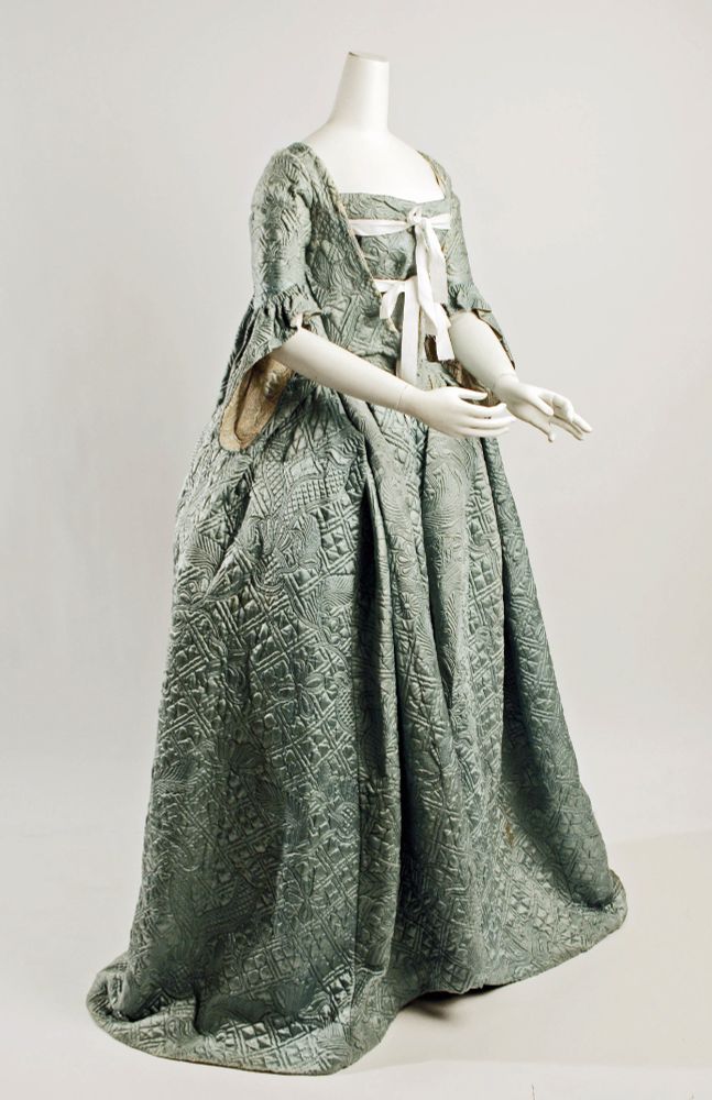 A photo of a mannequin wearing a quilted 18th century full length dress. Its made of silk quilting, with a sewn pattern of diamonds and flowers. The hips are quite wide, and the sleeves are elbow length with very wide hanging cuffs. The outer layer of the top is open at the front, tied over an inner layer with two broad white ribbons. 