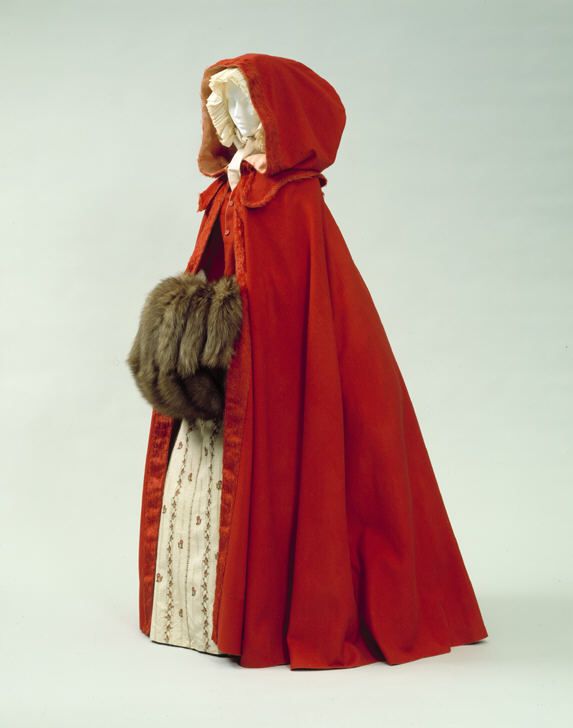Image 1. Mannequin wearing a full length red woolen cape with hood. The hood has been created extremely large at the rear to accommodate a headpiece or coiffured hair maybe. Its accompanied by (or made with) a matching red buttoned vest. The cape is completed by a small collar to the shoulders.