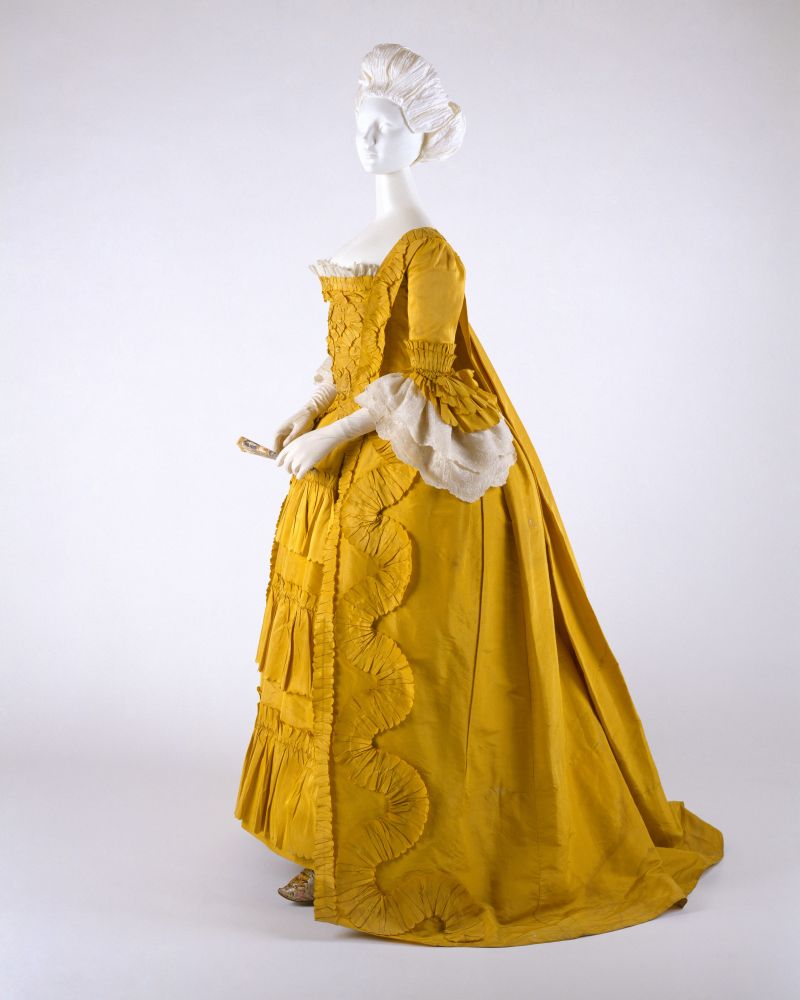 Image 1. Mannequin wearing a yellow court dress in the "Robe à la Française" style. The dress is full length. with a square neckline, elbow length sleeves, and a rear composed of voluminous folds extending into a slight train. The entire front of the dress has a lot of additional detailing (ruffles and other details that I don't have the terminology to describe), while the back is unadorned. The ends of the sleeves have both lace ruffles and ruffles in the same material as the dress. 