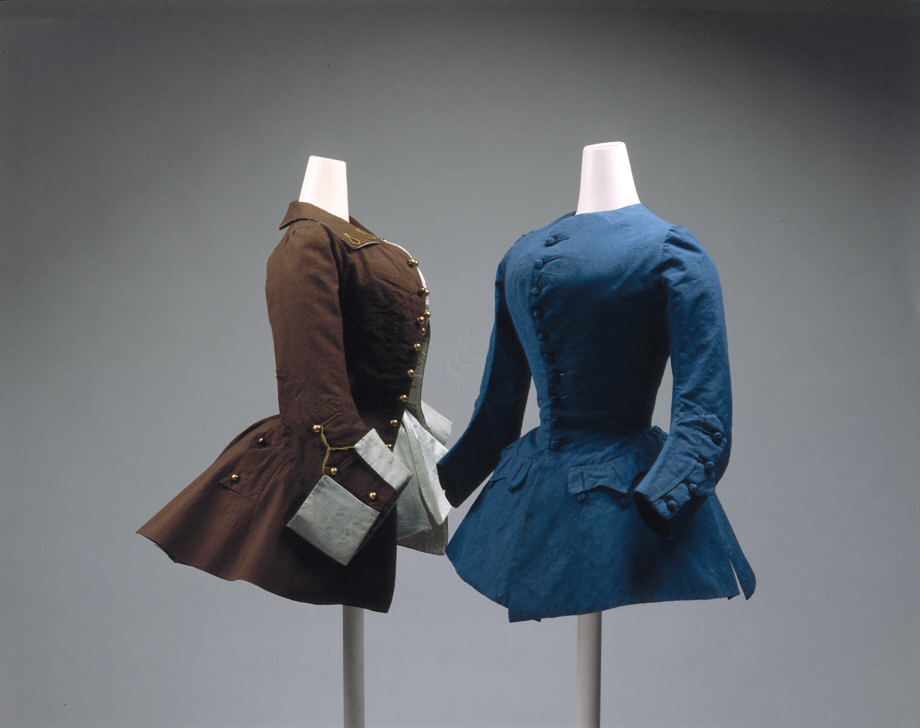 Two riding coats are shown in this picture.
Left - a Brown short riding coat, viewed from the side. It has a large collar, rounded buttons on the right lapel (possibly brass), and a short skirt that would probably just cover the hips when seated. It has oversized cuffs on the sleeves, turned back to show the white lining.
Right - a similar garment in blue. This one has no collar, and the buttons are the same colour as the main fabric. The cuffs on this garment are narrow. 