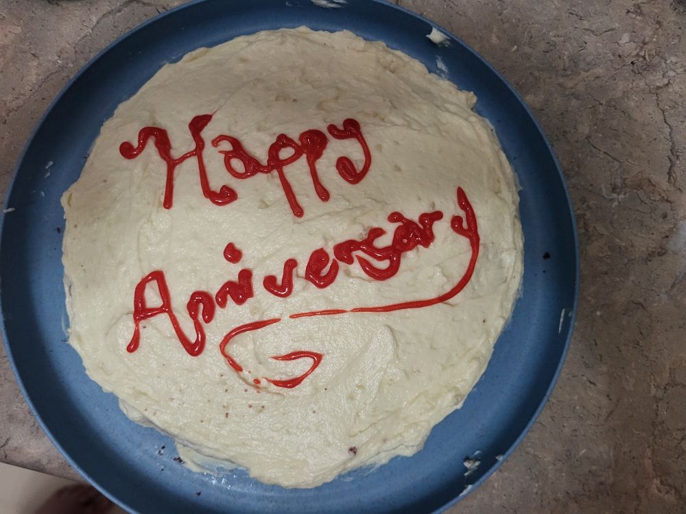 A simple white cake that's messily frosted with red glittery writing that says "Happy Anniversary". The writing is a bit sloppy but legible.