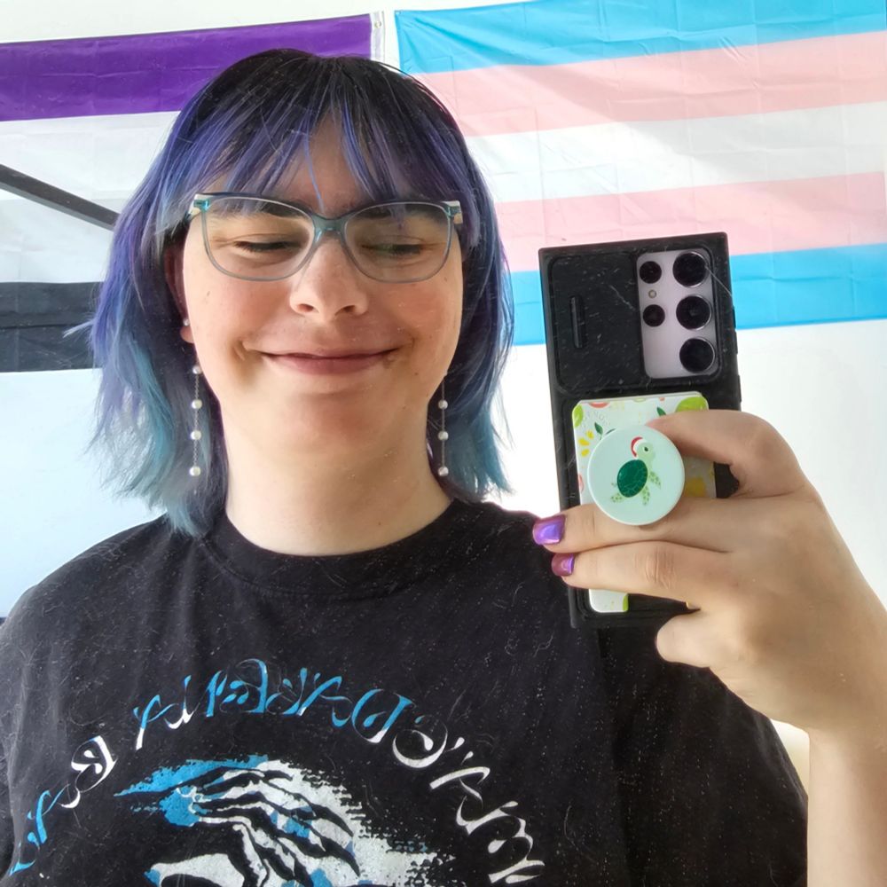 Girl with purple and blue hair and a black T-shirt with a design on it in long dangly pearl earrings in a bedroom with a bed behind her that has blue bedding and a ace pride flag and a trans pride flag behind her 