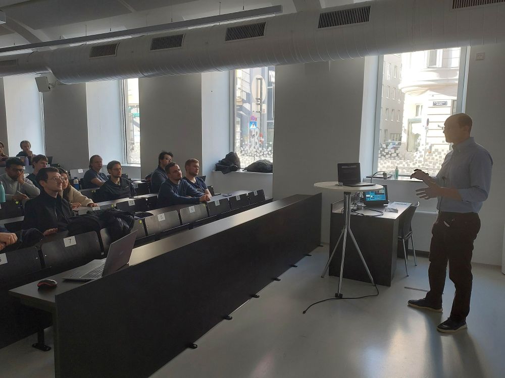 A lecturer is giving a presentation in a modern classroom with large windows. He is gesturing with his hands while speaking to an audience of students seated in tiered rows. The audience, consisting mostly of young adults, is attentively listening, some with laptops open.