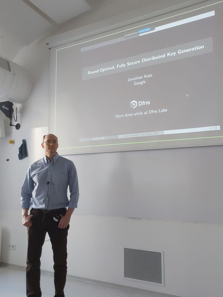 Jonathan Katz stands in a classroom beside a large screen, which displays the title slide of a presentation titled "Round-Optimal, Fully Secure Distributed Key Generation." The slide also states that the work was done at Dfns Labs. He is attentively looking towards the audience.
