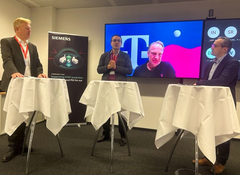 This image shows a panel discussion, featuring four men. Three men are physically present, standing behind podiums with white covers, and one man is participating virtually, displayed on a large screen in the background. The setting includes a Siemens banner, emphasizing cybersecurity. The physical participants are attentively listening to the speaker, who is gesturing as he speaks. 