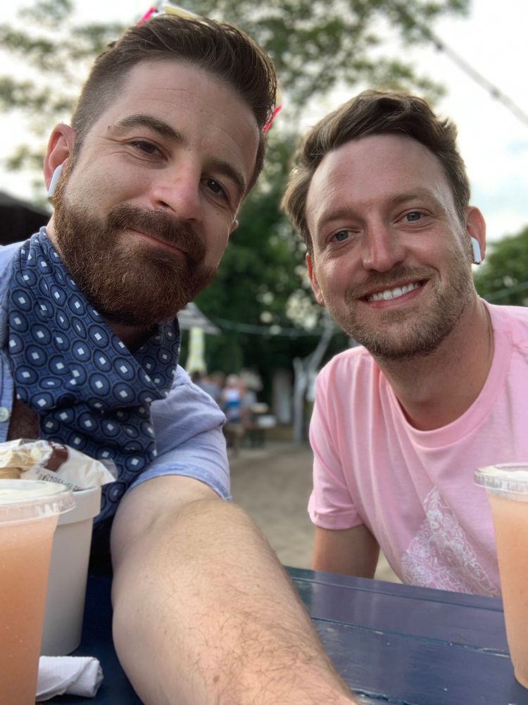 This is a selfie of my husband and me in Provincetown. The phone is on a picnic table and shows two gays in short sleeve shirts, one has a neck bandanna because of course he does.