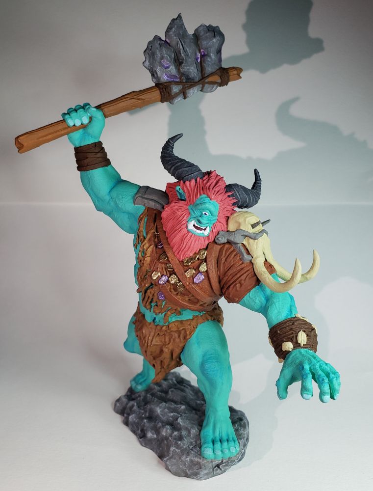 The Borborygmos MTG mini. It is a very large cyclops, brandishing a stone axe with a wooden handle above its head. He has a large mane of bright red hair and a red beard, and blue-ish green skin. Two large, dark grey horns shoot out from his head, and he is wearing leather bands on his wrists, and some pelts for clothing. His mouth is open, two large tusks protruding from the bottom row of teeth. On his left, the skull of an elephant is his shoulder-plate. 