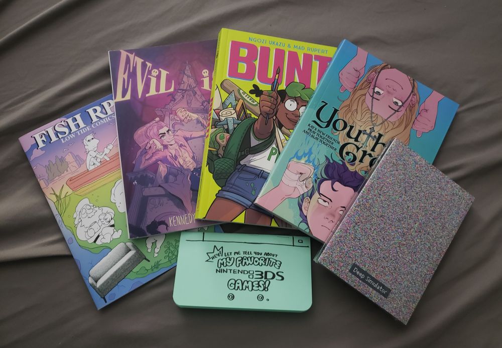5 books rest in an arc with 1 zine sitting in the gap below it. From left to right, the books are Fish RPG: Low Tide Comics, Evilish, Bunt!, Youth Group, and Deep Simulator. The small blue zine below is titled "Hey! Let Me Tell You About My Favorite Nintendo 3DS Games!"
