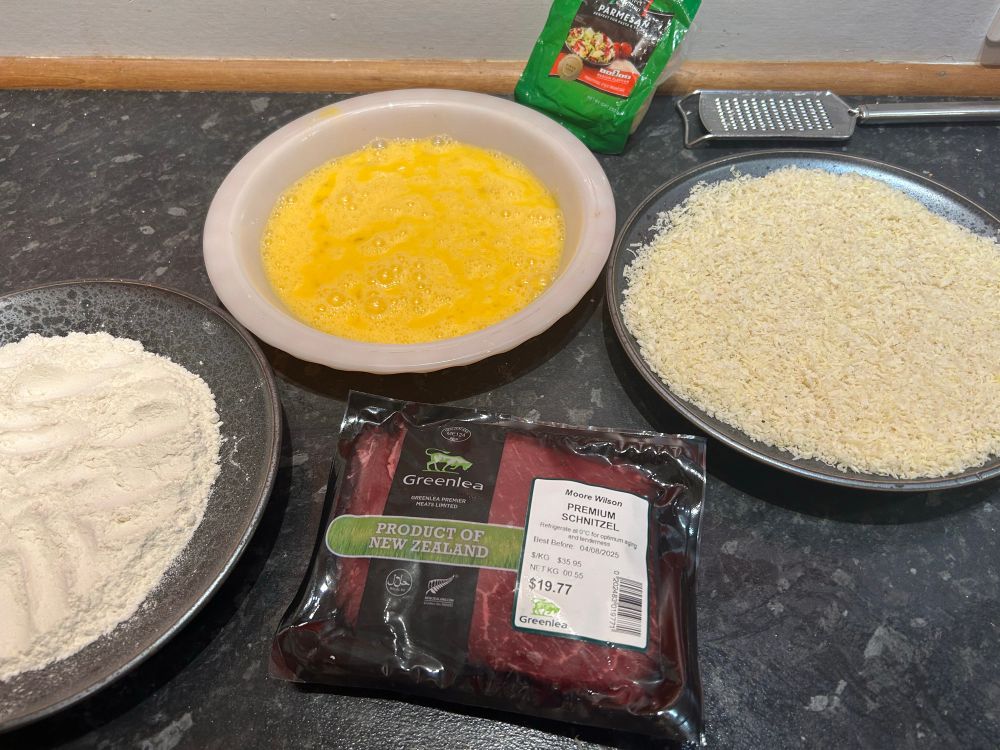 Plates of flour, egg and panko breadcrumbs ready to crumb the schnitzel which is in a packet in front. 