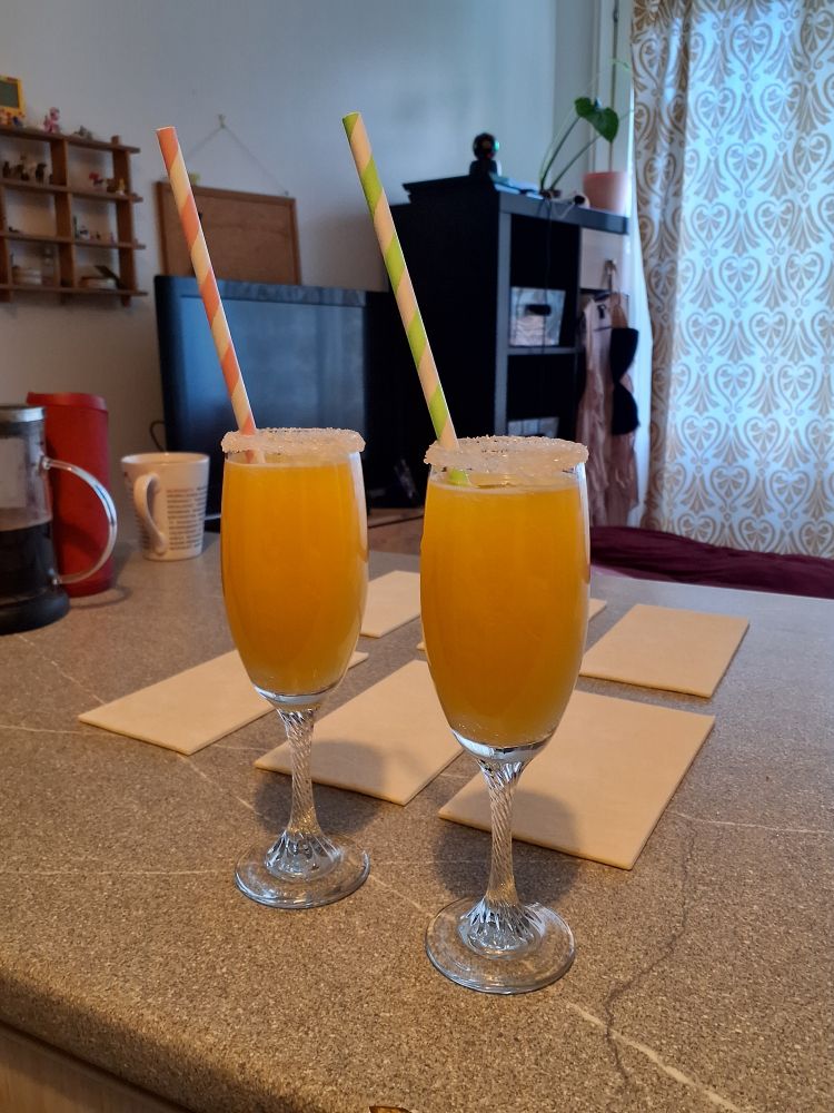 Kaksi mimosaa sokerireunustetuissa kuohuviinilaseissa. Toisessa vihreä ja toisessa vaaleanpunainen paperipilli. Taka-alalla sulamassa lehtitaikinalevyjä.