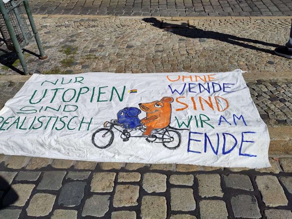 Auf dem Boden liegendes Banner mit Elefant und Maus auf einem Fahrrad. Der Text lautet 

Nur Utopien sind realistisch 
Ohne Wende sind wir am Ende 