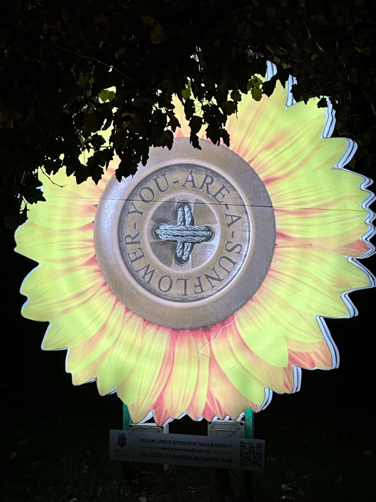 Künstlicher Blütenkopf einer Sonnenblume, ca. 1,5m hoch, auf Sockel in Park bei Nacht. Gelbe Blütenblätter außen, die Mitte sieht aus wie ein Knopf. Kreisrunder Schriftzug am Knopf: „You are a sunflower“