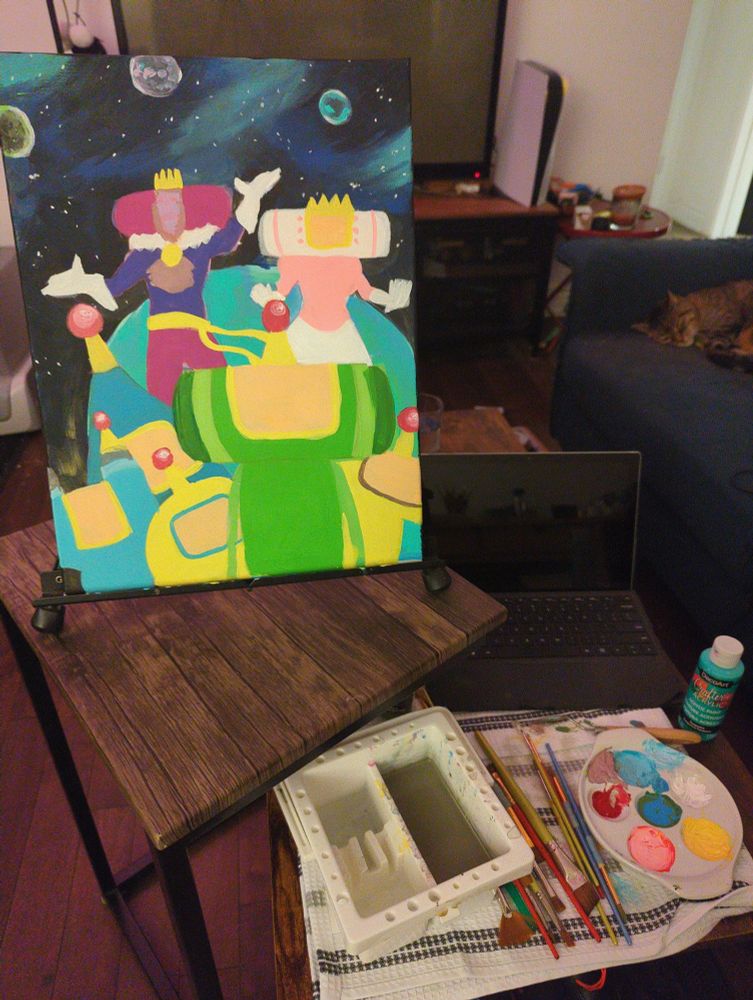 a photograph of my painting set up. my canvas is on a small easil that's sitting on a side table. there's a half-finished painting of katamari damacy fanart I've been working on, done in acrylic and gouache. on my coffee table is my brushes, pallet, paints, and laptop (for reference photo). my roommates cat is asleep on the couch in the top corner of the photograph.