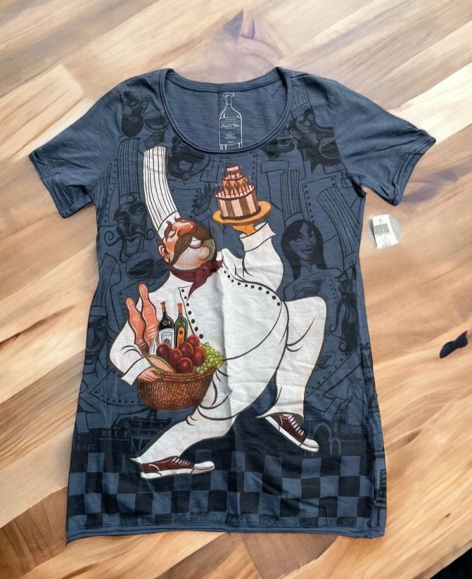 A dark grey woman's shirt with a giant chef on it. He's marchine with a cake AND a basket of fruits wines and french breads. He's also wearing a dope pair of chucks. There are a bunch of other chefs in the background in black on grey giving him the side eye. 
