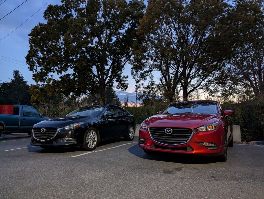 A picture of a black Mazda3 parked next to a red Mazda3. Both cars are from the same generation and possibly the same year.