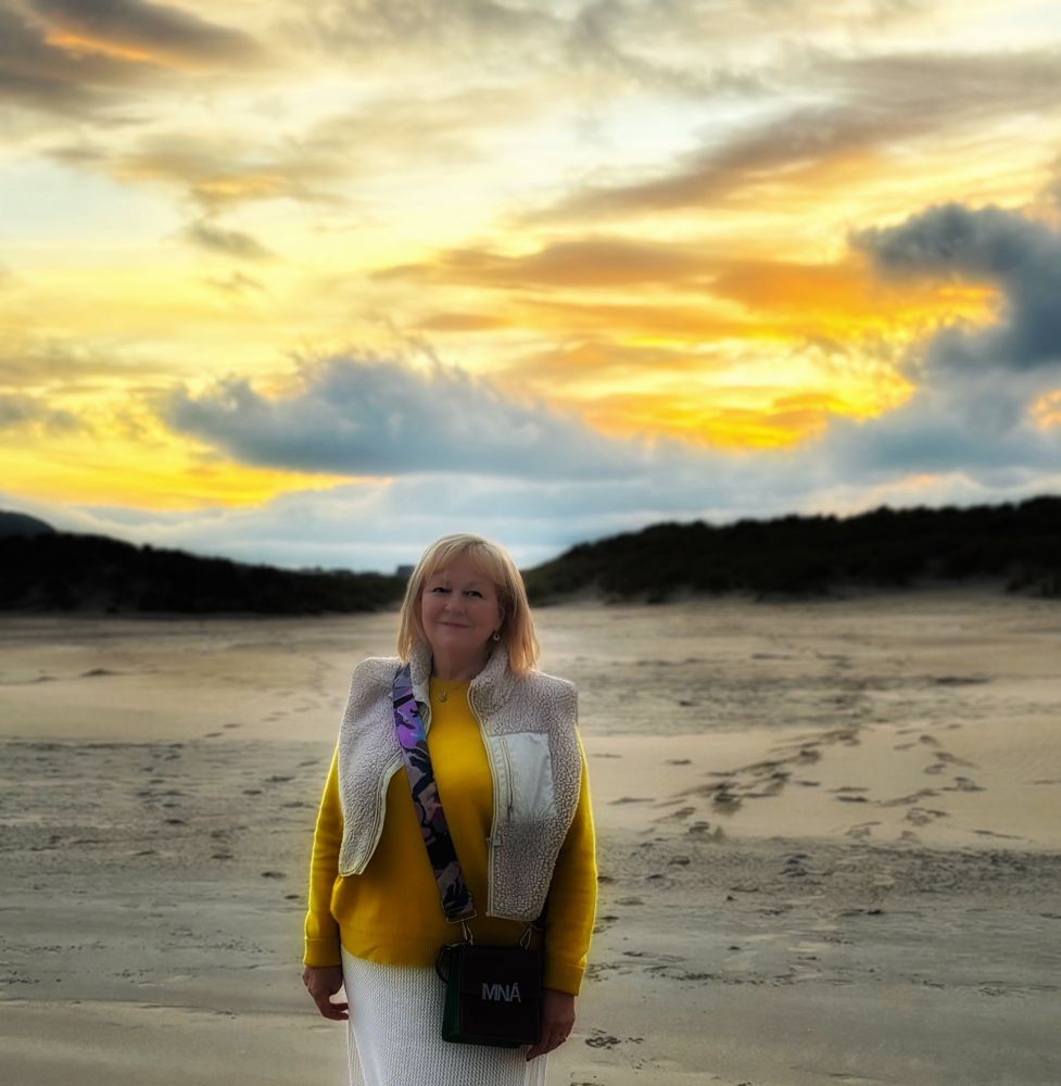 Me pictured against a gorgeous orange and golden yellow sunset. I’m standing on a sandy beach. I’m wearing a yellow sweater and cream skirt and a fleece waistcoat with a brown bag slung over my shoulder. I’ve got blonde collar length hair and a smile 😊 