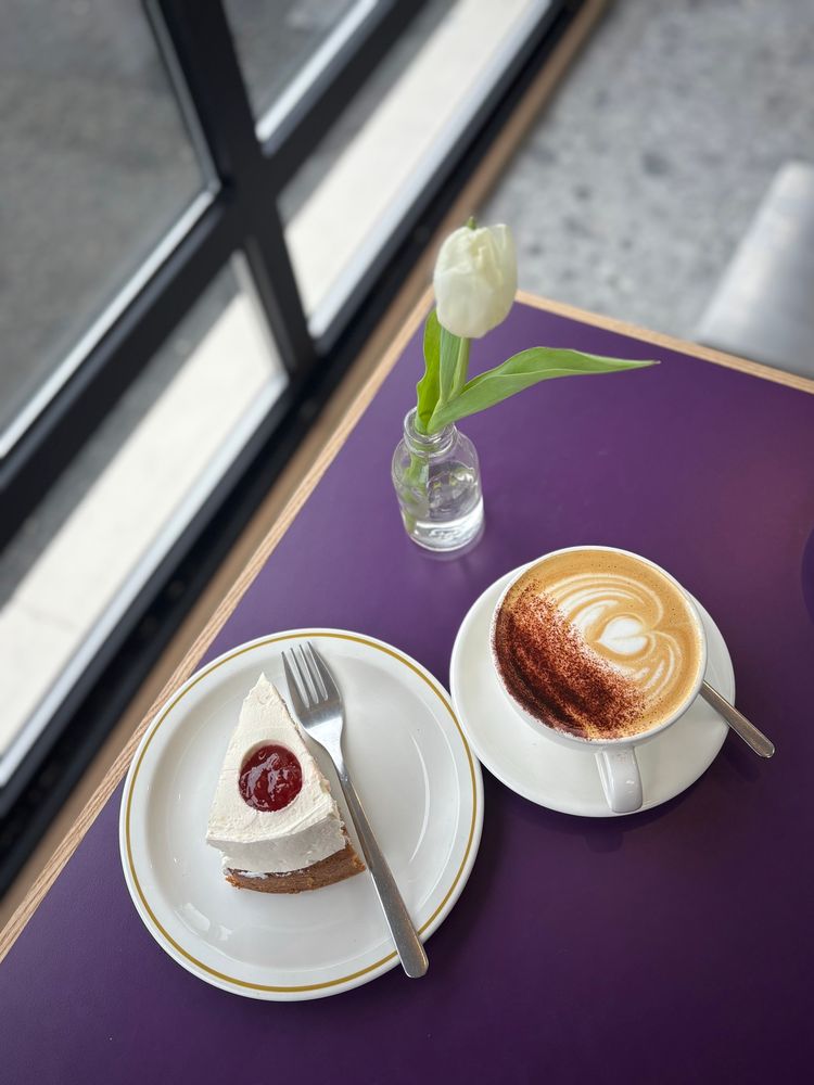A cappuccino and a slice of sponge cake topped with jam & buttercream. White crockery on a purple table by a window with a single white tulip in a vase