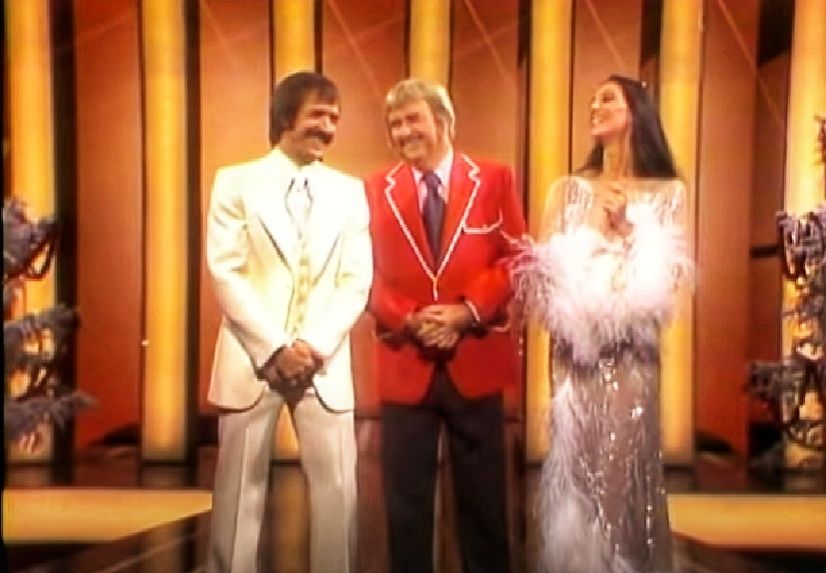 Sonny Bono on the left in a white 3 piece suit. Captain Kangaroo Bob Keeshon in the center with a red jacket & dark pants, cher to the right in a silver white sequined dress with feather boa sleeve cuffs. From 1976 Christmas special
 