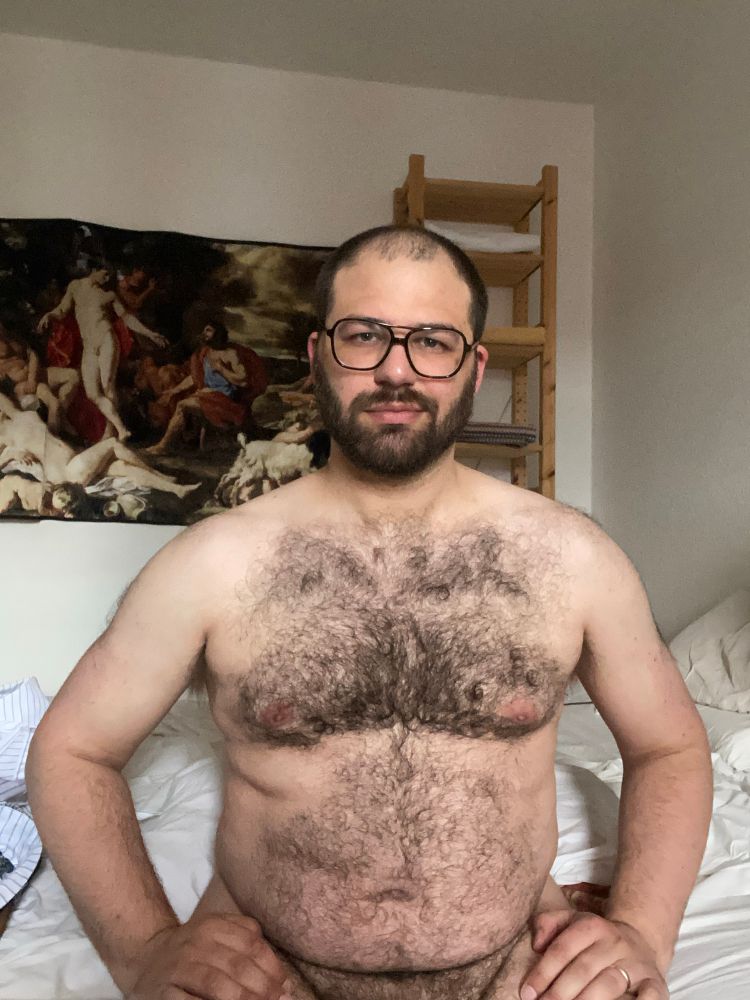 Naked cubby sitting on the bed, he has a fresh haircut and beard trim. On the wall behind him is a print of Poussin’s Midas and Bacchus