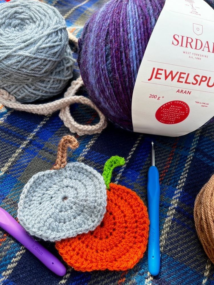 A photo of assorted coloured yarn, with two crocheted pumpkin coasters and two crochet hooks next to them.