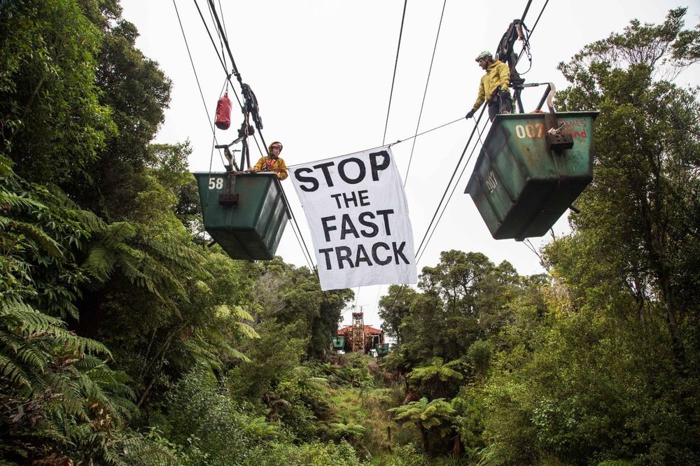 Two people wearing hard hats and jackets stand in coal cars that are high up in the air on a coal track, surrounded by beautiful bush. A banner hangs between them saying Stop the Fast Track
