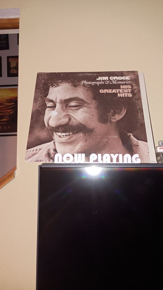 I vinyl record album displayed in a now playing sign. Jim Croce's Greatest hits. Cover is a black and white photograph of Jim croce. 
