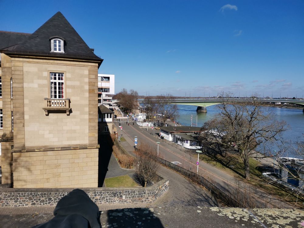 Blick vom Alter Zoll in Bonn, Richtung Kennedybrücke. Es ist sonnig, einige Leute spazieren die Rheinpromenade entlang. Links im Vordergrund das historische Institut der Uni Bonn, eer Kopf des Fotographen wirft einen Schatten geradeaus