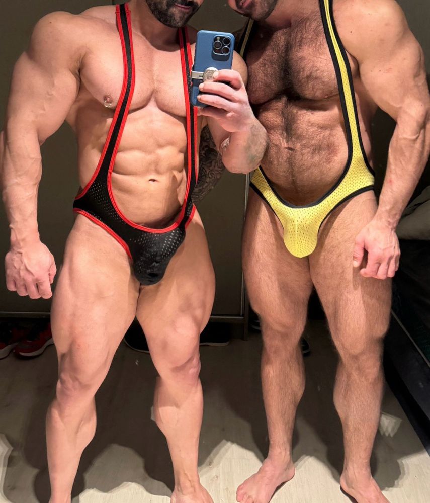 The image show a gay couple wearing wrestling singlets. The one on the left has a bodybuilder physique with his erect penis bulging through the black and red singlet. The one on the right is also muscular, but with a lot of body hair, and his big balls and penis are also bulging through his yellow singlet.