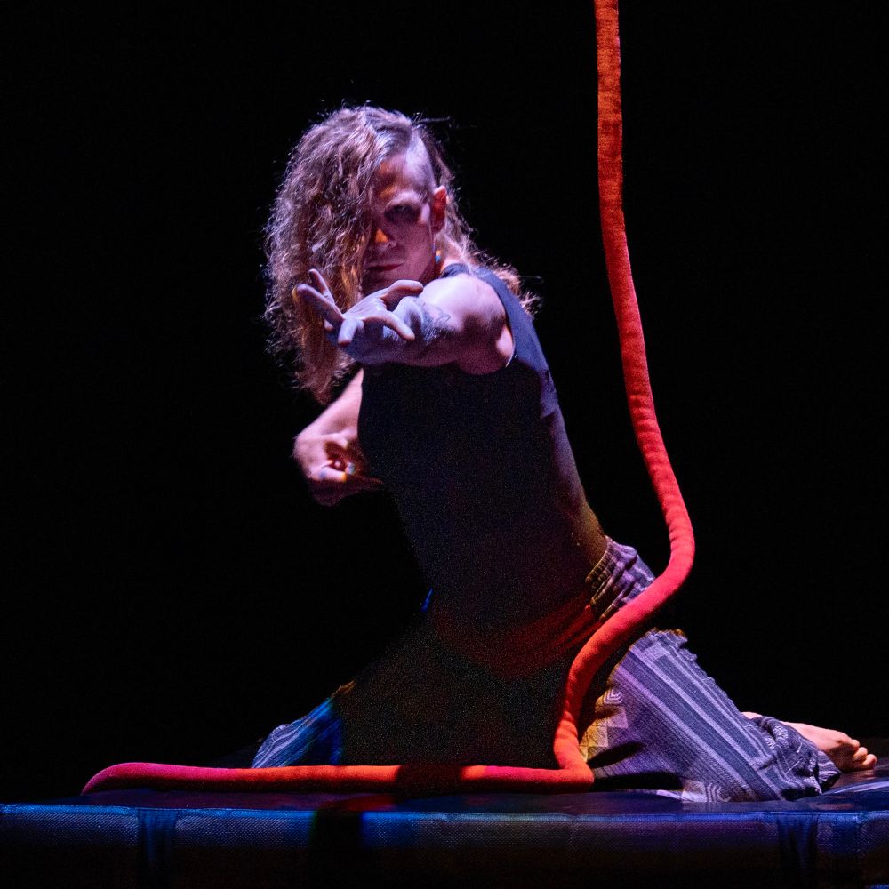 An aerial rope artist, kneeling on the floor. Their arm is stretched out in front of them and they are looking intently at the camera. A red aerial rope hangs down beside them and is draped over their thigh and coiled on the floor.