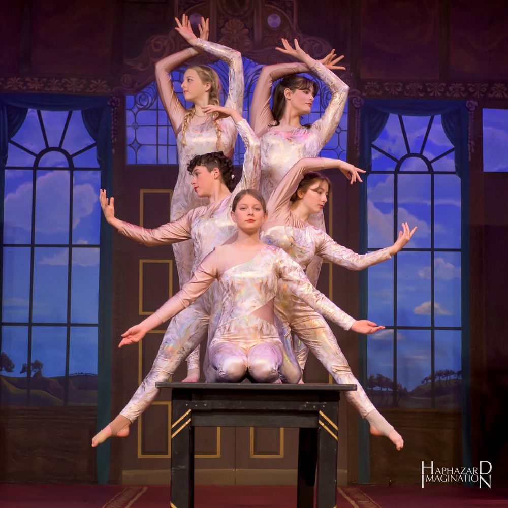 5 circus artists in a group pose on a table. They are wearing white shimmery body outfits with their arms and legs in artistic shapes, not quite symmetrical. The backdrop shows a painting of ornate doors and windows with a quiet landscape and sky with a few clouds.