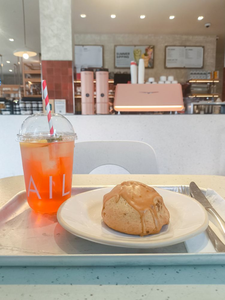 A plastic café takeaway cup of bright orange iced tea next to an iced scone on a small white plate. Both are on a metal tray on a table in front of the barista station, which has a giant, pastel pink fancy-looking coffee machine on it front & centre.