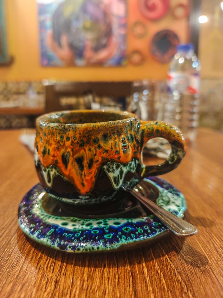 A small, handcrafted cup and saucer with a coffee spoon. The crockery is bubbled and looks like it was dipped in deep ochre and navy paint. The cup contains Arabic coffee, a spiced coffee prepared similarly to Turkish coffee.
