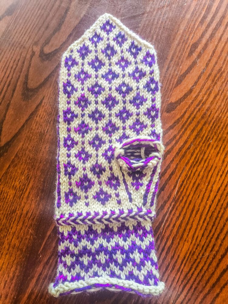 The almost complete back of a partially-knitted mitten knit in white wool as the base colour and variegated purple-magenta wool as the contrast colour. The thumb still needs to knit, leaving a hole in the palm. The pattern on the wrist is three rows of purple diamonds, with a slightly larger middle row that has white crosses in the middle of the diamonds. Above the wrist is a Latvian braid, a knitting technique that produces a raised, horizontal stripe in alternating colours (it looks similar to a fishtail hair braid). The main body of the mitten is made up of a repeating pattern of diamonds with white centres with single contrast-colour stitches between them.
