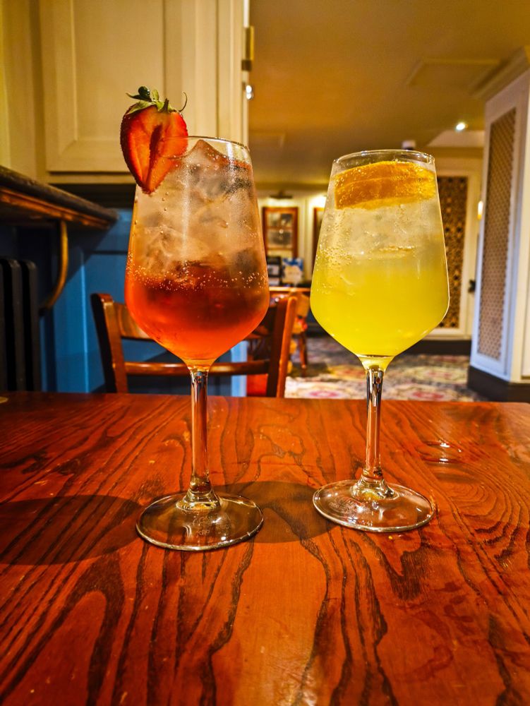 Two cocktails on a small, dark wooden table, both in wine glasses. The left cocktail is deep red in the bottom third of the glass and the rest is filled with a pale pink clear drink, with half a strawberry stuck on the rim. This is a peach blush spritz: rosé wine, peach schnapps, lemonade. The right cocktail is bright, slight cloudy yellow in the bottom half, topped up with clear, very pale yellow fizzy drink with a couple of lemon slices stuck in the top of the drink. This is a limoncello spritz: limoncello, prosecco, soda water.