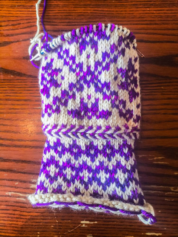 The front of a partially-knitted mitten knit in white wool as the base colour and variegated purple-magenta wool as the contrast colour. The pattern on the wrist is three rows of purple diamonds, with a slightly larger middle row that has white crosses in the middle of the diamonds. Above the wrist is a Latvian braid, a knitting technique that produces a raised, horizontal stripe in alternating colours (it looks similar to a fishtail hair braid). The main body of the mitten is made up of several large diagonally-oriented squares pieced together with eight-pointed stars or snowflakes (I'm not quite sure which they're supposed to be) inside.