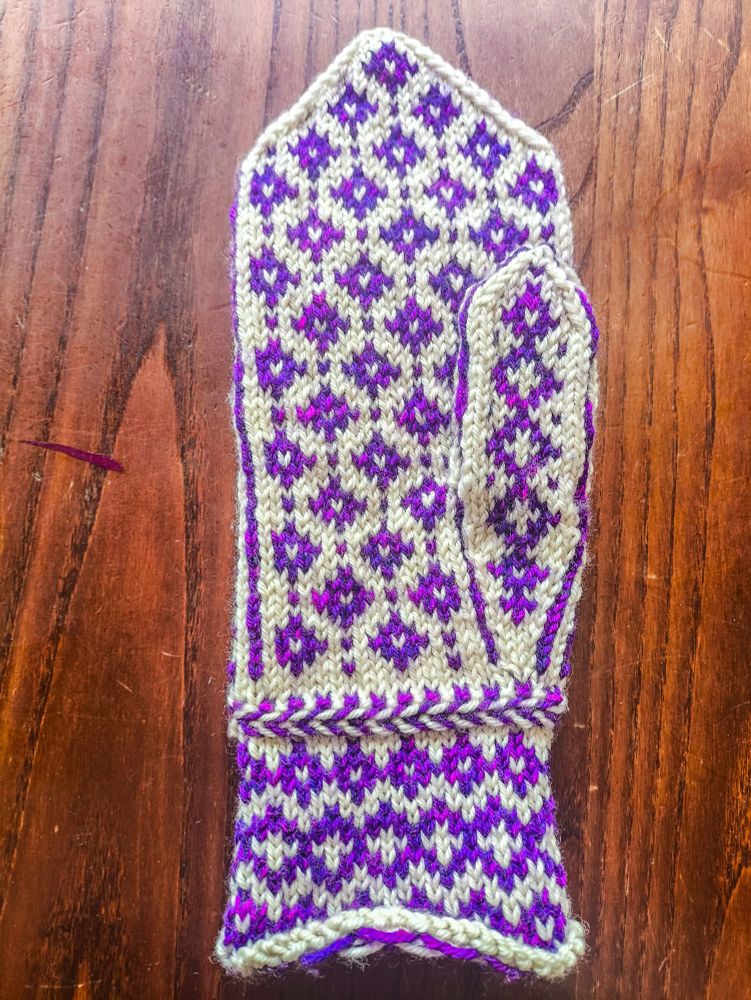 The back of a complete mitten knit in white wool as the base colour and variegated purple-magenta wool as the contrast colour. The pattern on the wrist is three rows of purple diamonds, with a slightly larger middle row that has white crosses in the middle of the diamonds. Above the wrist is a Latvian braid, a knitting technique that produces a raised, horizontal stripe in alternating colours (it looks similar to a fishtail hair braid). The main body of the mitten is made up of a repeating pattern of diamonds with white centres with single contrast-colour stitches between them. The thumb has a vertical line of larger diamonds with white crosses in the centre. 
