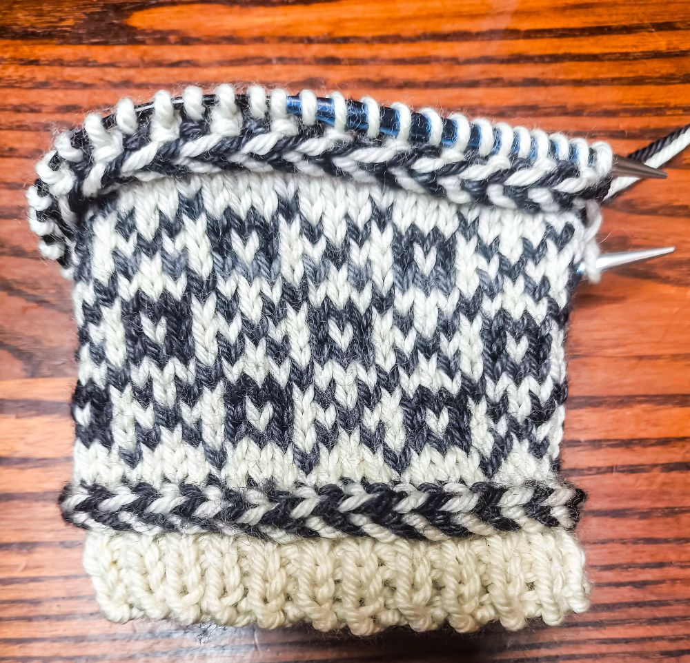 The cuff of a mitten-in-progress, knit in white wool as the base colour and semi-solid black/charcoal grey wool as the contrast colour. From the bottom up: a small section of 1x1 rib, then a Latvian braid (a knitting technique that carries the yarn in front and twists it around itself to create horizontal "braids" in your knitting), a colourwork section made up of squares with white centres in the middle of diamonds created by a large crosshatch pattern, then another Latvian braid. 