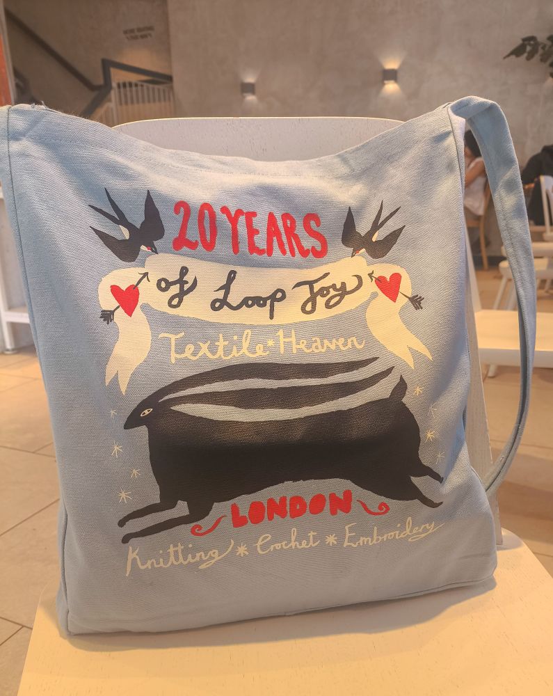 A large sky blue tote bag with a long strap propped up on a chair in my local Gail's. All the text looks handwritten, but in slightly different fonts. At the top, in red it says “20 years". Below that, on a banner with two lovehearts pierced by arrows and held up by a swallow at each side, it says "of Loop Joy". Below that are the words "Textile Heaven". Below that is a large black, stylised rabbit. Below the rabbit in red block capitals it says "London", then below that "Knitting * Crochet * Embroidery".