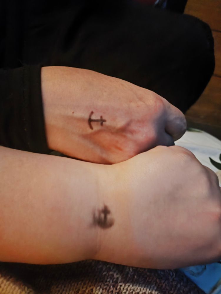 Two pale hands next to each other. Both have a small anchor ink-stamp stamped on them. The anchor is in the centre of the upper hand and is closer to the wrist and is smudged on the lower hand (my hand).