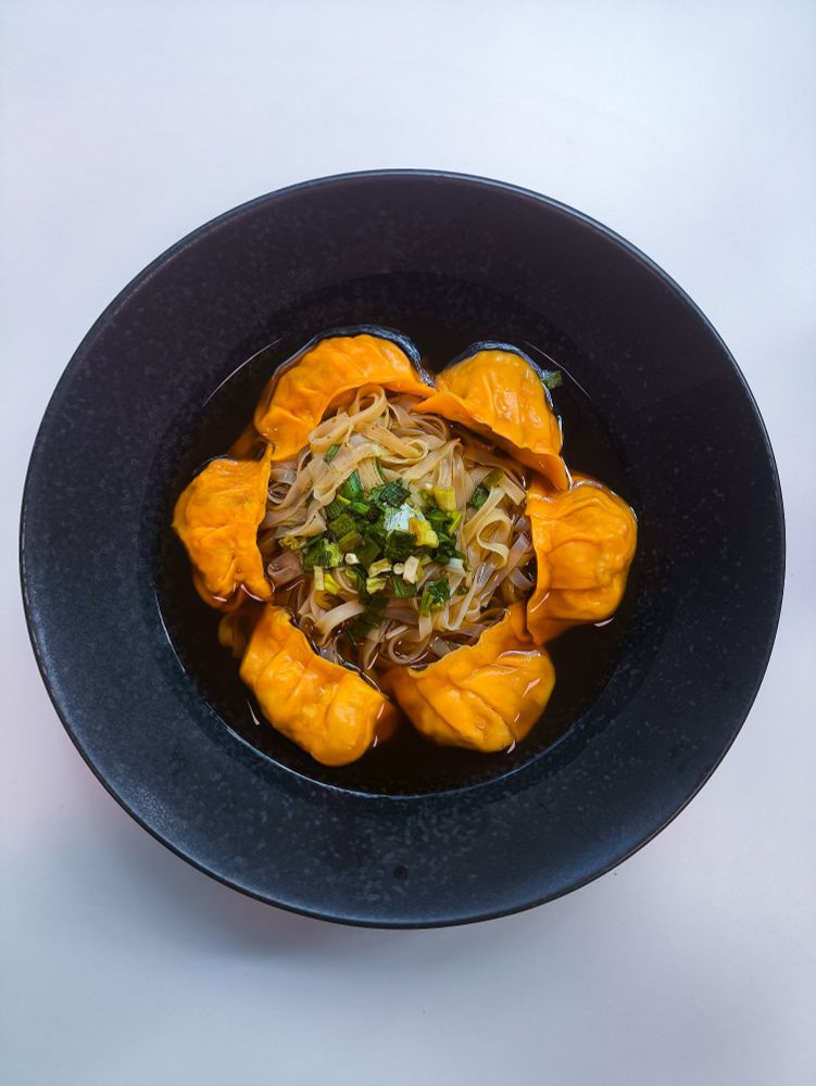 A black crockery ramen bowl on a white desk. It contains a pile of rice noodles with spring onion pieces on top, surrounded by bright orange king prawn gyoza, topped off with a dark soy sauce broth.