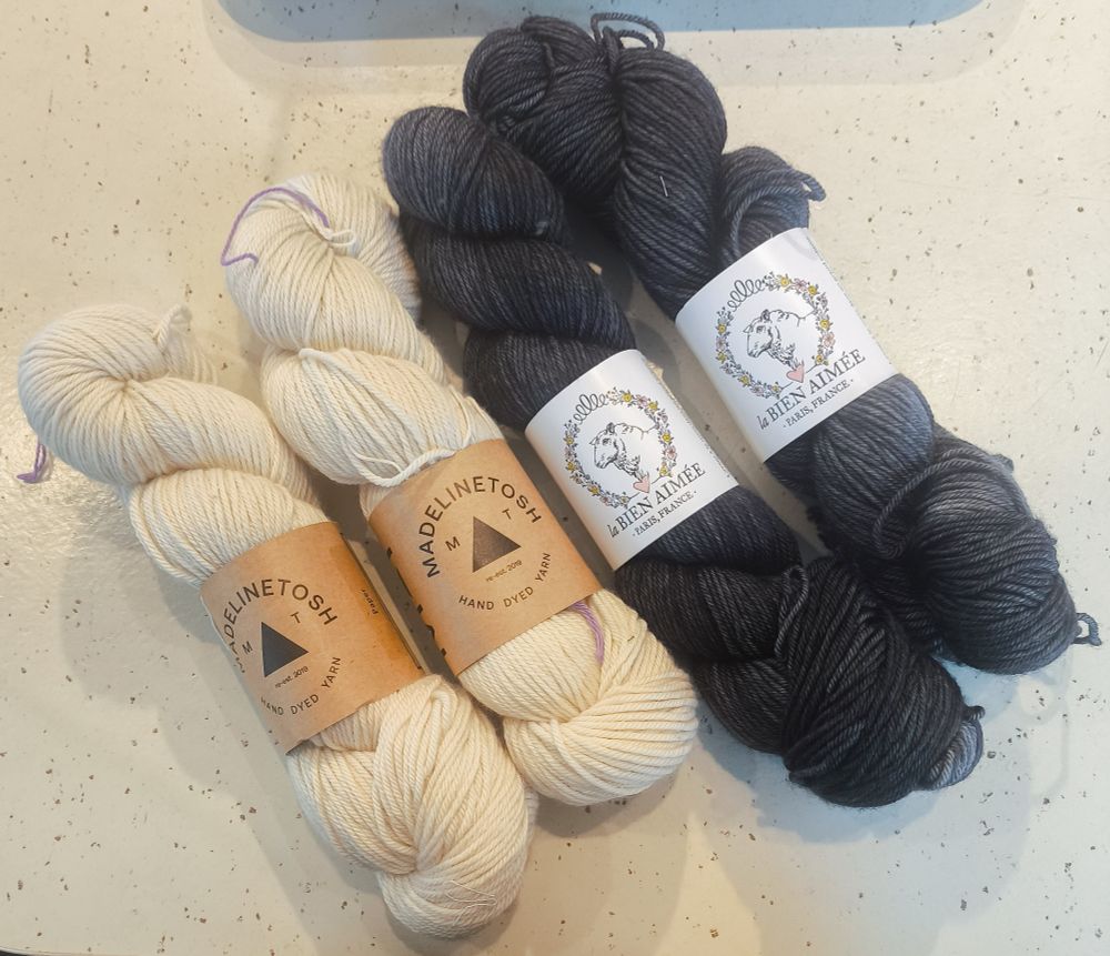 Four skeins of yarn laid out on a white table with brown speckles. Two left two skeins are off-white with a brown label saying "Madelinetosh hand dyed yarns" in a circle around a black triangle. The right two skeins are longer and thinner, and are semi-solid black, like a yarn version of faded black jeans. These yarns have a white label with a drawing of a sheep's head in a circle of flowers and foliage, with a pink loveheart and the words "la Bien Aimée Paris, France" underneath.